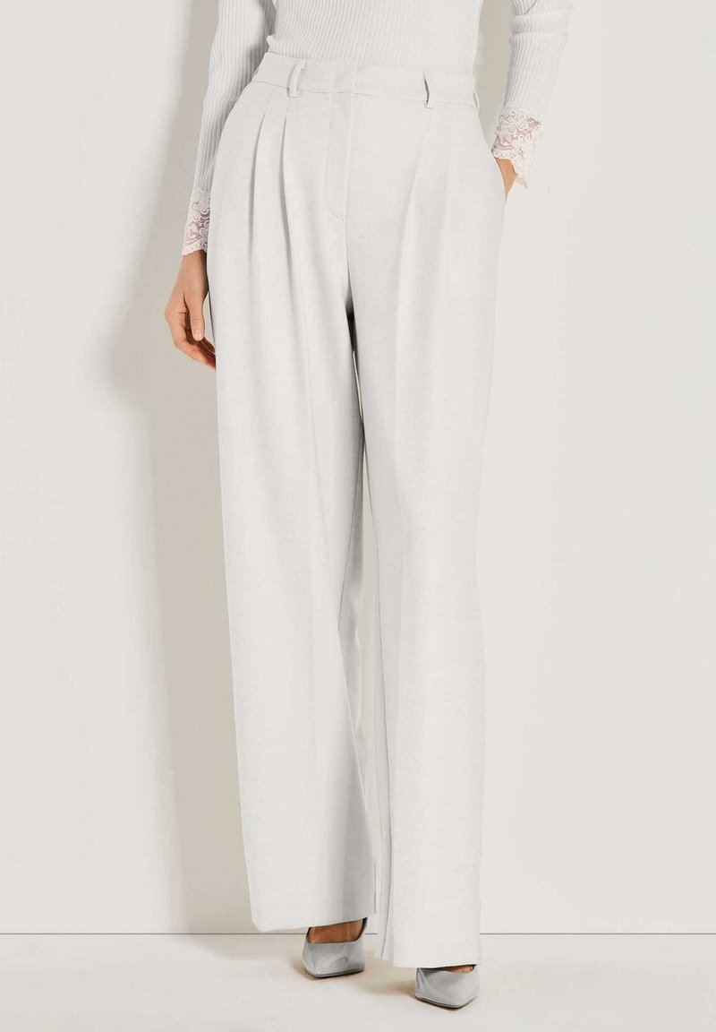 Vrouw in hoge taille, wijde witte broek gecombineerd met een witte geribbelde trui met kantenkraag en zilveren puntige hakken.