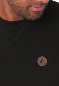 Černá mikina s žebrovaným límcem, křížkovým zdobením a kulatou hnědou nášivkou s logem na hrudi. Viditelná měkká textura látky.