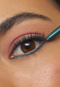 Teal eyeliner påføres den nedre øyelokket; har brune øyne med definerte vipper og myk ferskenøyenskygge. Huden viser fine teksturer.