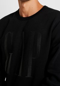 Sweat-shirt noir avec un grand logo "GAP" en relief dans une teinte plus foncée. Design classique à col rond avec un tissu doux et texturé.