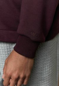 Burgunderfarbener Sweatshirtbund mit geripptem Textur, versehen mit geprägt eingraviertem Branding. Die Hand zeigt Details des Stoffs und eine Nahaufnahme der Designelemente.