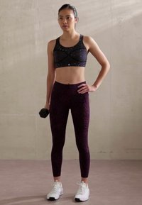 Violet raštuoti leggingai ir juodas sportinis liemenukas su kryžminiu dizainu; laiko hantelį, vilkėdama baltus sportinius batus.