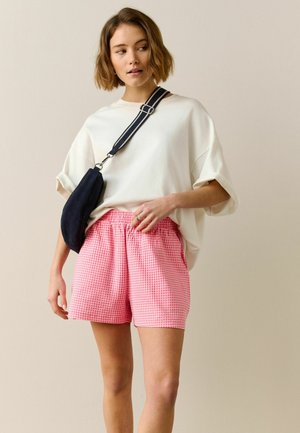 Woman wearing loose white T-shirt, pink checkered shorts, and black crossbody bag with striped strap, standing against plain background.