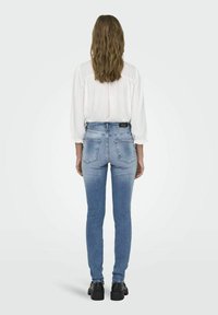 Jeans de tiro alto en azul claro con un ajuste slim, ligeramente desgastados, y dos bolsillos traseros, combinados con una blusa blanca y zapatos negros.