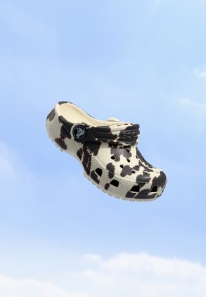 Un sabot à imprimé vache crème et brun flotte sur un ciel bleu parsemé de nuages légers.