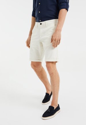 Homme portant un short blanc à ourlet roulé, une chemise bleu marine à boutons et des mocassins noirs sans lacets, debout devant un fond blanc.
