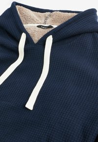 Sweat à capuche bleu marine avec un extérieur en tricot gaufré texturé et des cordons de serrage crème ; doublé d'une douce polaire beige à l'intérieur.