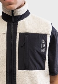 Gilet en polaire avec une partie supérieure de couleur crème et un panneau inférieur noir, doté d'une poche zippée et d'un logo de marque imprimé sur la poitrine.