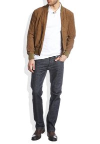 Hombre vistiendo una chaqueta bomber de ante marrón, camiseta polo blanca, jeans oscuros y botas de cuero marrón oscuro, de pie con una mano en el bolsillo.