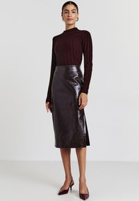 Top a maniche lunghe a coste color bordeaux abbinato a una gonna a tubino lucida in pelle marrone scuro a vita alta con uno spacchetto laterale. Scarpe con tacco completano il look.