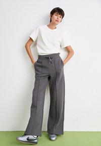 Femme se tenant contre un mur blanc, portant un t-shirt blanc, un pantalon large gris avec taille ajustable et des baskets argentées.