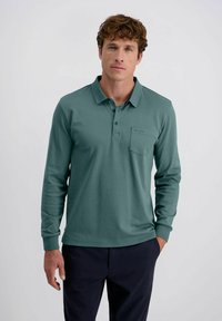 Mann mit lockigem Haar, bekleidet mit einem türkisfarbenen Langarm-Polohemd und dunklen Hosen, steht mit einer Hand in der Tasche vor einem schlichten Hintergrund.