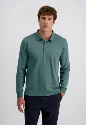 Man with curly hair wearing a teal long-sleeve polo shirt and dark pants, standing with one hand in pocket against a plain background.