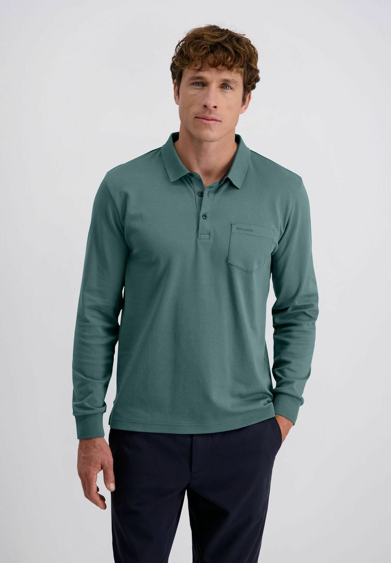 Mann mit lockigem Haar, bekleidet mit einem türkisfarbenen Langarm-Polohemd und dunklen Hosen, steht mit einer Hand in der Tasche vor einem schlichten Hintergrund.