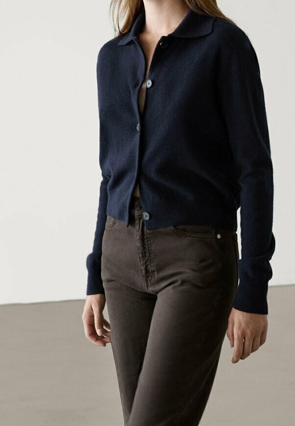Femme portant un cardigan bleu marine boutonné et un pantalon taille haute marron foncé, debout devant un fond clair uni.