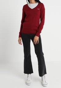 Röd v-ringad tröja, stickad struktur, bärs över en grå t-shirt. Matchad med svarta utsvängda jeans och vita sneakers, stående mot en neutral bakgrund.