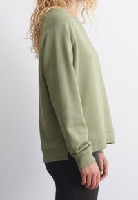 Zelená mikina s dlouhými rukávy, žebrovanými manžetami a kulatým výstřihem. Hladká textura, volný střih a minimalistický design bez viditelných vzorů.