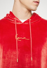 Sudadera de terciopelo rojo con un logotipo bordado en oro. Presenta una capucha con cordón, una textura suave y acentos metálicos en los extremos del cordón.