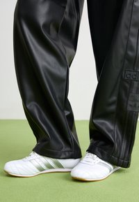 Zwarte sweatpants van nep leer met een losse pasvorm, voorzien van een Adidas-logo patch en zwarte verticale strepen, gecombineerd met witte schoenen en zilveren accenten.