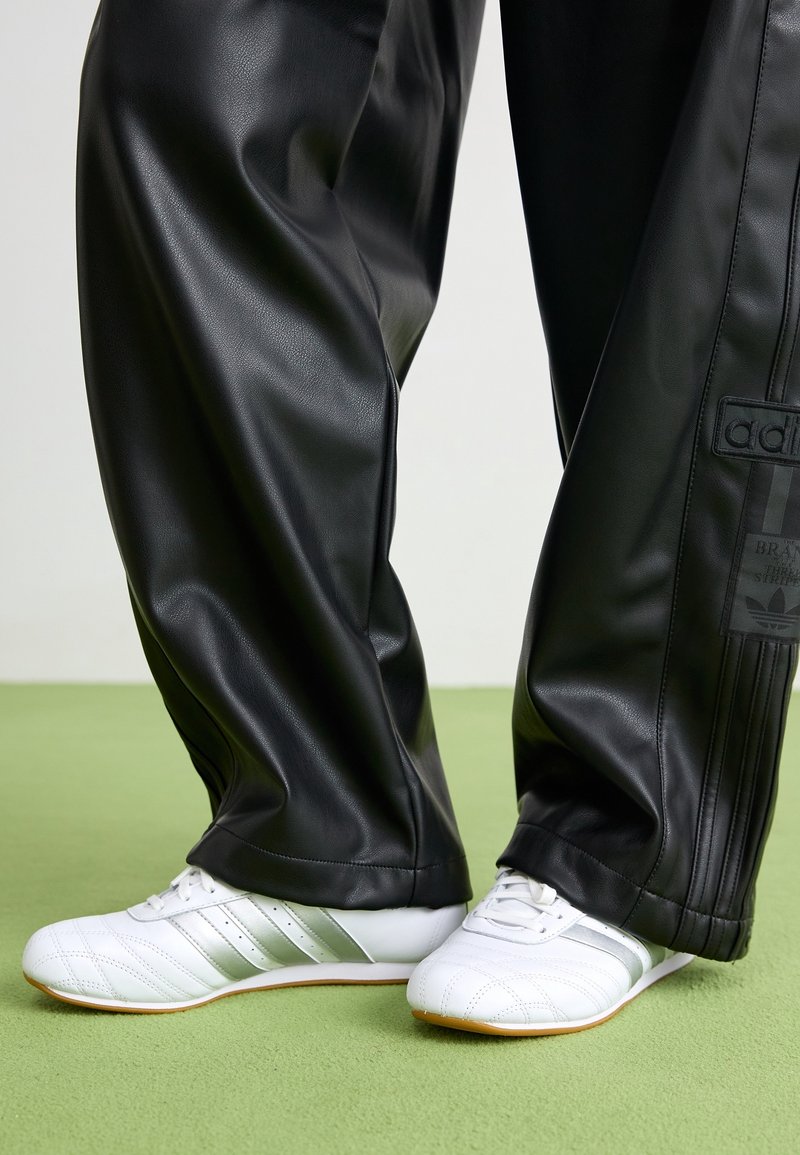 Zwarte sweatpants van nep leer met een losse pasvorm, voorzien van een Adidas-logo patch en zwarte verticale strepen, gecombineerd met witte schoenen en zilveren accenten.