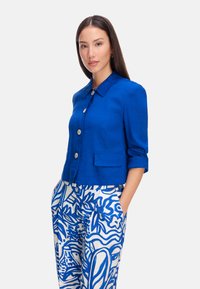 Blue linen crop jacket with a pointed collar, front buttons, and a pocket. Paired with patterned blue and white trousers featuring floral designs.