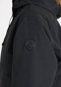 Svart vattentät jacka med en texturerad yta, med en rund logotypapplikation på ärmen. Designen inkluderar en huva och justerbara snoddar.