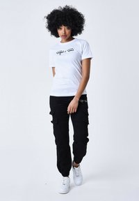 T-shirt bianca in cotone con stampa grafica nera, abbinata a pantaloni cargo neri con tasche e polsini elasticizzati, indossati con scarpe da ginnastica bianche.
