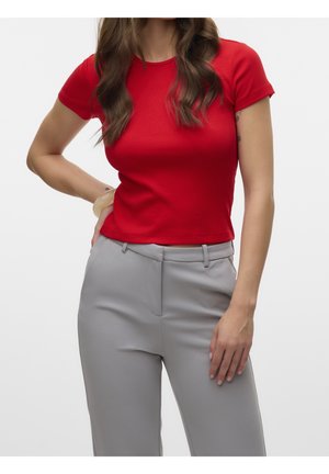 Femme portant un t-shirt rouge ajusté à manches courtes et un pantalon gris clair taille haute, une main sur la hanche et l'autre reposant sur le côté.