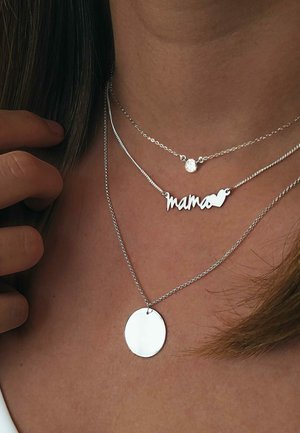 Drei schichtweise angelegte Silberneklaces um einen Hals, darunter ein kleiner Edelstein, eine "Mama" Namensplatte mit einem Herz und ein runder Anhänger.