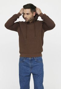 Brauner Strickpullover mit Kapuze, strukturiertem Oberteil, langen Ärmeln und verstellbaren Kordeln. Getragen mit blauen Jeans.