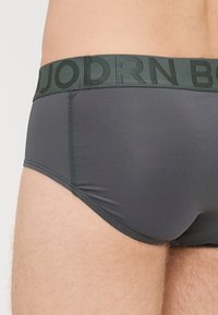 Pilkas apatinis trikotažas su tekstūruotu paviršiumi ir žaliu juosmeniu, kuriame iškalti raidės. Siuvimo detalės palei kraštus. Formos prigludimas.