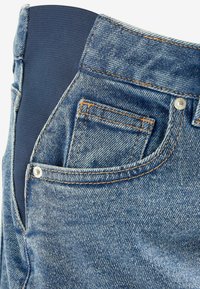 Jeans de mezclilla con una cinturilla elástica oscura, lavado azul claro, costuras naranjas y un bolsillo delantero con un botón de presión plateado.