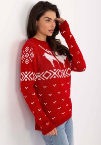 Frau in rotem Weihnachtspullover mit weißen Rentier- und Geometriemustern, die mit der Hand an der Stirn posiert und eine hellblaue Jeans trägt.