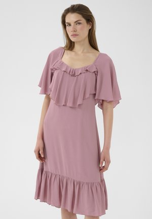 Kobieta w różowej, zakurzonej sukience midi z falującymi rękawami, marszczonym dekoltem i marszczonym dołem, stojąca na jednolitym białym tle.