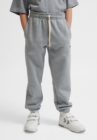 Grijze sweatpants met een elastische tailleband en verstelbaar trekkoord, voorzien van zijzakken en geribbelde boorden. Gecombineerd met witte sneakers.