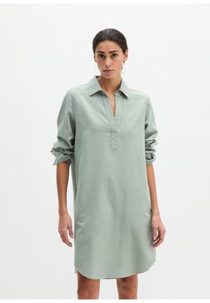 Femme portant une robe chemise ample vert clair à manches longues avec col et encolure en V, manches retroussées, debout devant un fond blanc uni.