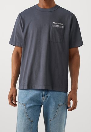 Homme portant un T-shirt Abercrombie gris foncé avec poche poitrine et un jean en denim bleu clair, se tenant devant un fond uni.