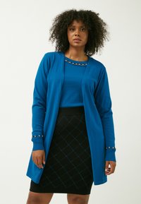 Cardigan lungo blu sopra una maglietta blu e una gonna nera. Il cardigan ha dettagli dorati sulle maniche. La gonna presenta un motivo a diamante strutturato.