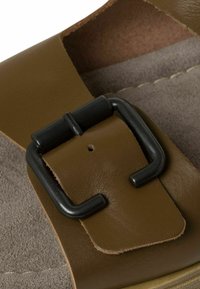 Sandale en cuir marron avec une large sangle, boucle en métal noir et semelle intérieure douce et texturée. Couture contrastante visible le long des bords.