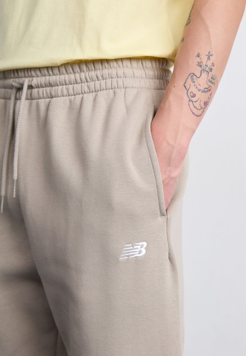 Helle graue Jogginghosen mit elastischem Bund, seitlichen Taschen und weißem gesticktem Logo am linken Bein. Aus weichem Baumwollmaterial, mit lockerer Passform.