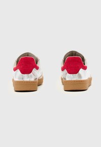 Fehér bőrből készült, piros velúr díszítéssel, gumitalpú tornacipő varrott részletekkel. Kerekded formájú és párnázott belsővel rendelkezik.