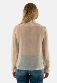 Blusa ligera de color beige claro, con cuello volante, mangas largas y puños elásticos. La tela tiene una textura ligeramente arrugada.