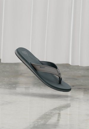 Szaro-czarna klapka marki Reef unosząca się nad polerowaną betonową podłogą z białymi zasłonami w tle.