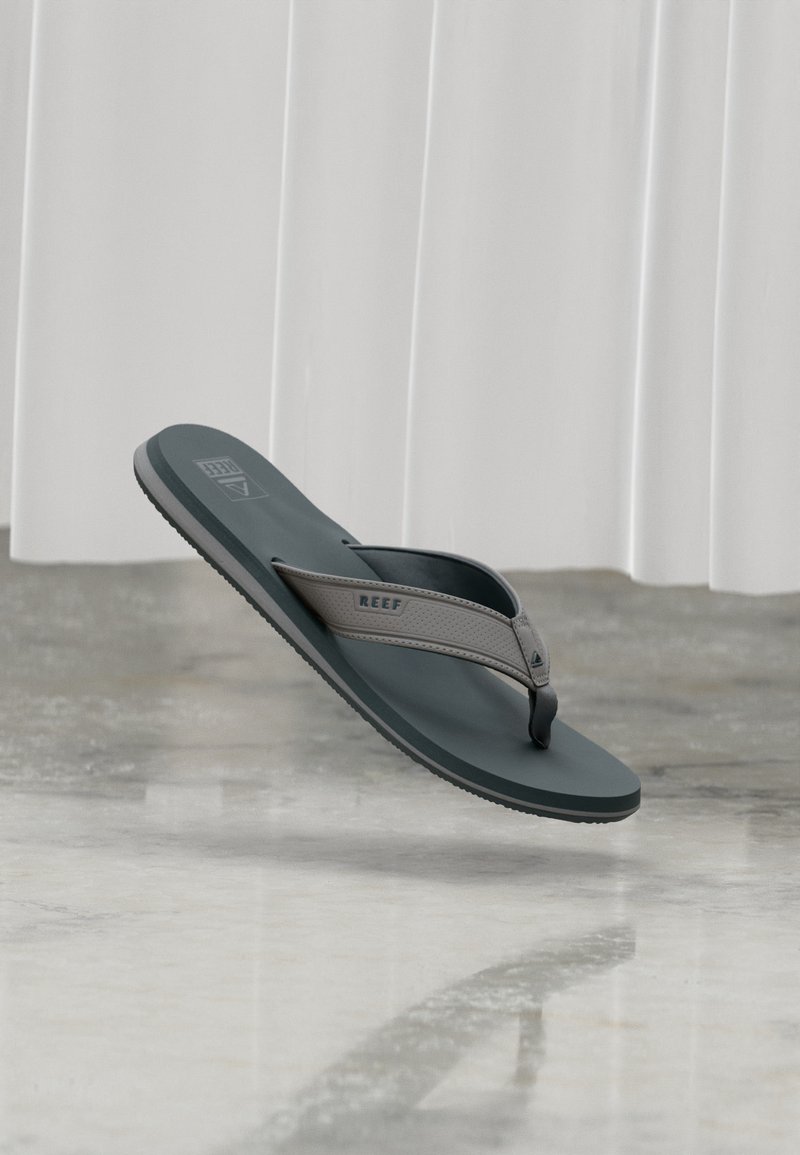 Γκρι και μαύρο σαγιονάρι Reef που αιωρείται πάνω από ένα γυαλισμένο δάπεδο από σκυρόδεμα με λευκές κουρτίνες στο φόντο.