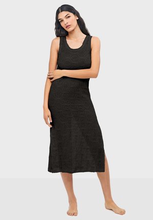 Mujer descalza de pie, vestida con un vestido midi negro sin mangas con un patrón geométrico texturizado, posando con un brazo cruzado sobre su cuerpo.