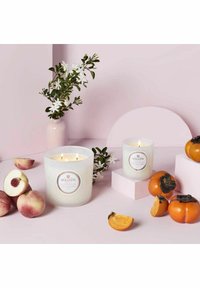 Voluspa-ljus i texturerat vitt glas, omgivna av persiko- och persimmonfrukter, på en mjuk rosa bakgrund med blommiga inslag.