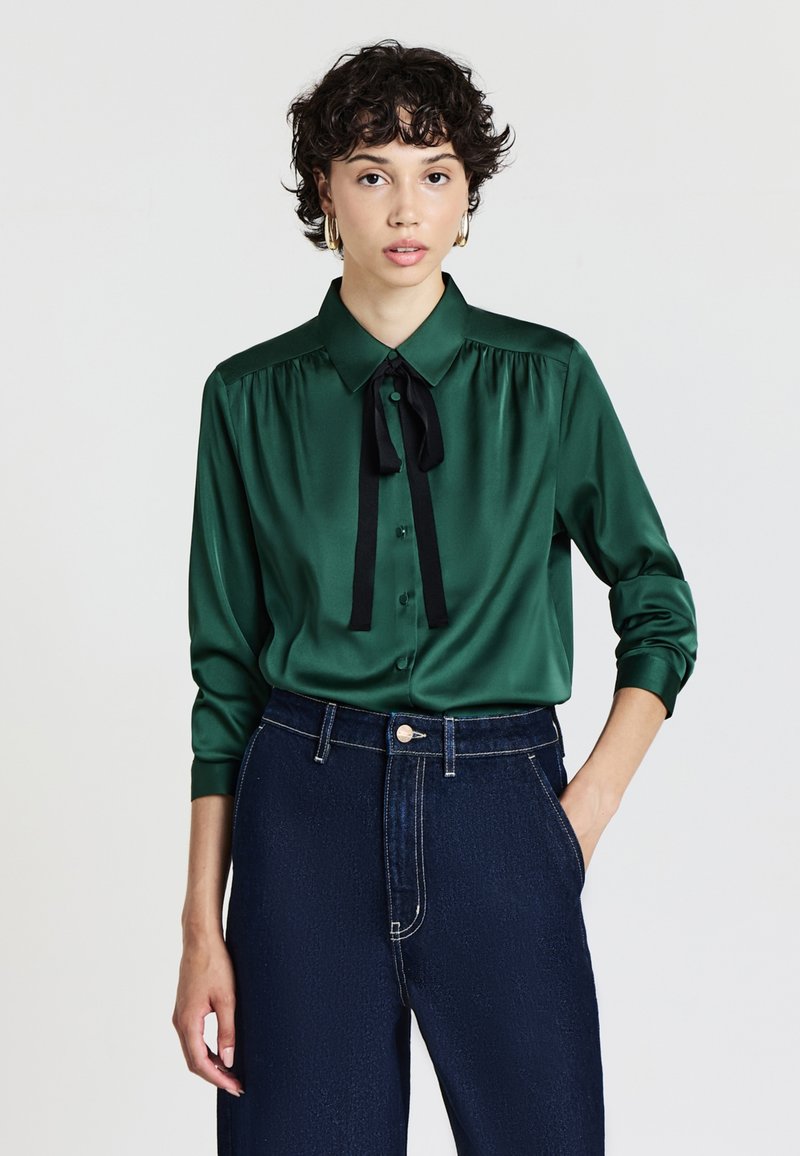 Blusa in satin verde con colletto classico, fiocco nero, chiusura con bottoni e maniche lunghe. Abbinata a pantaloni in denim blu a vita alta.
