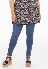 Femme portant un jean skinny bleu, un tunique à motif papillon coloré, et des sandales à talons compensés noires avec des semelles en liège, se tenant devant un fond blanc.