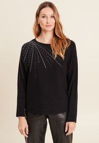 Schwarzer Pullover mit rundem Halsausschnitt und langen Ärmeln, verziert mit silbernen Nieten, die in einem strahlenförmigen Muster über die Schultern angeordnet sind.