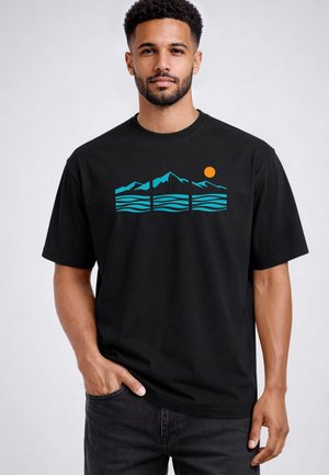 Mann trägt schwarzes T-Shirt mit türkisfarbenem Berg- und Wellendesign und kleinem orangefarbenem Sonnenmotiv, steht mit einer Hand in der Tasche vor einfachem Hintergrund.